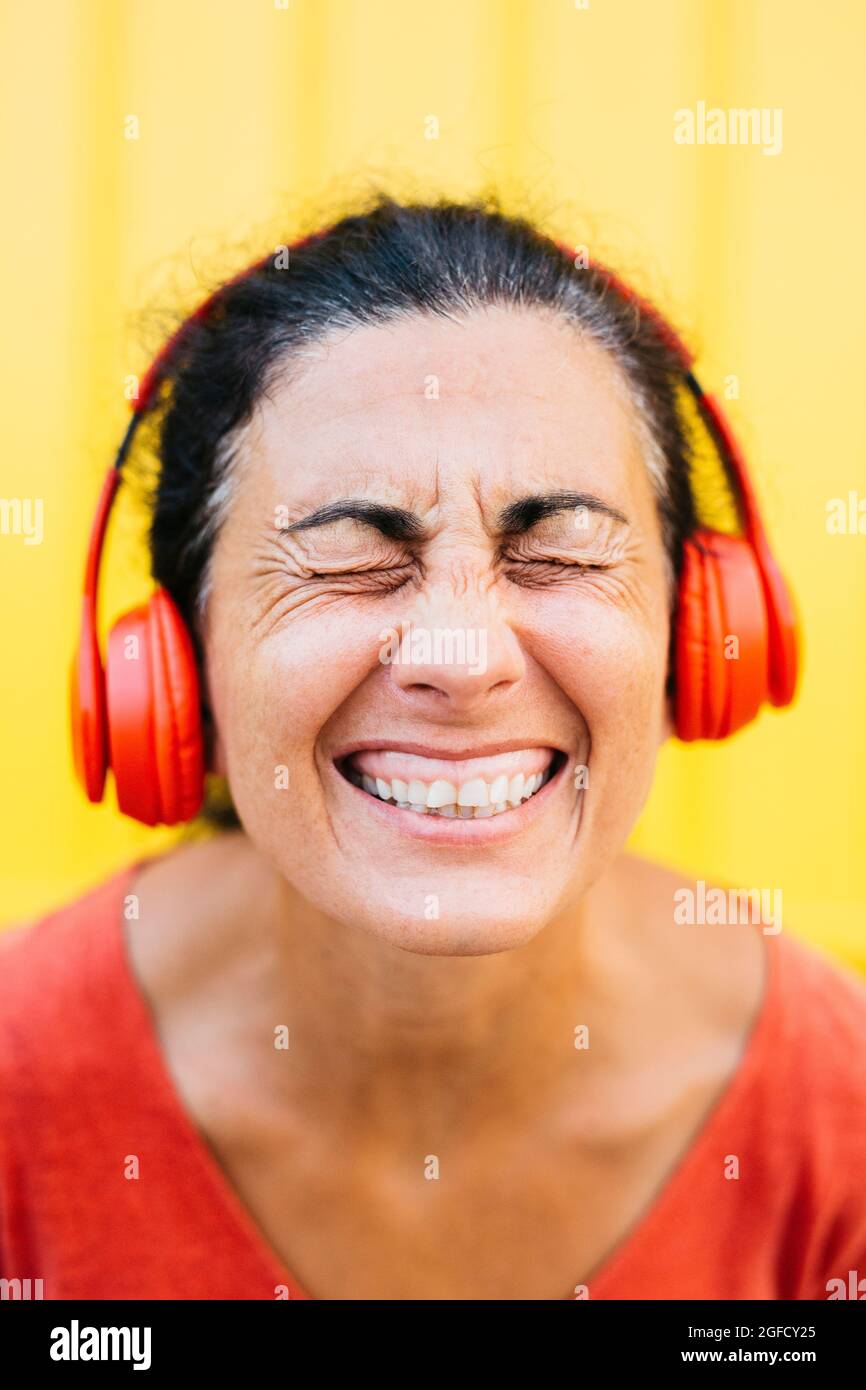 Donna di mezza età che indossa abiti rossi, ascolta musica con uno smartphone rosso e una cuffia wireless rossa, su sfondo giallo Foto Stock