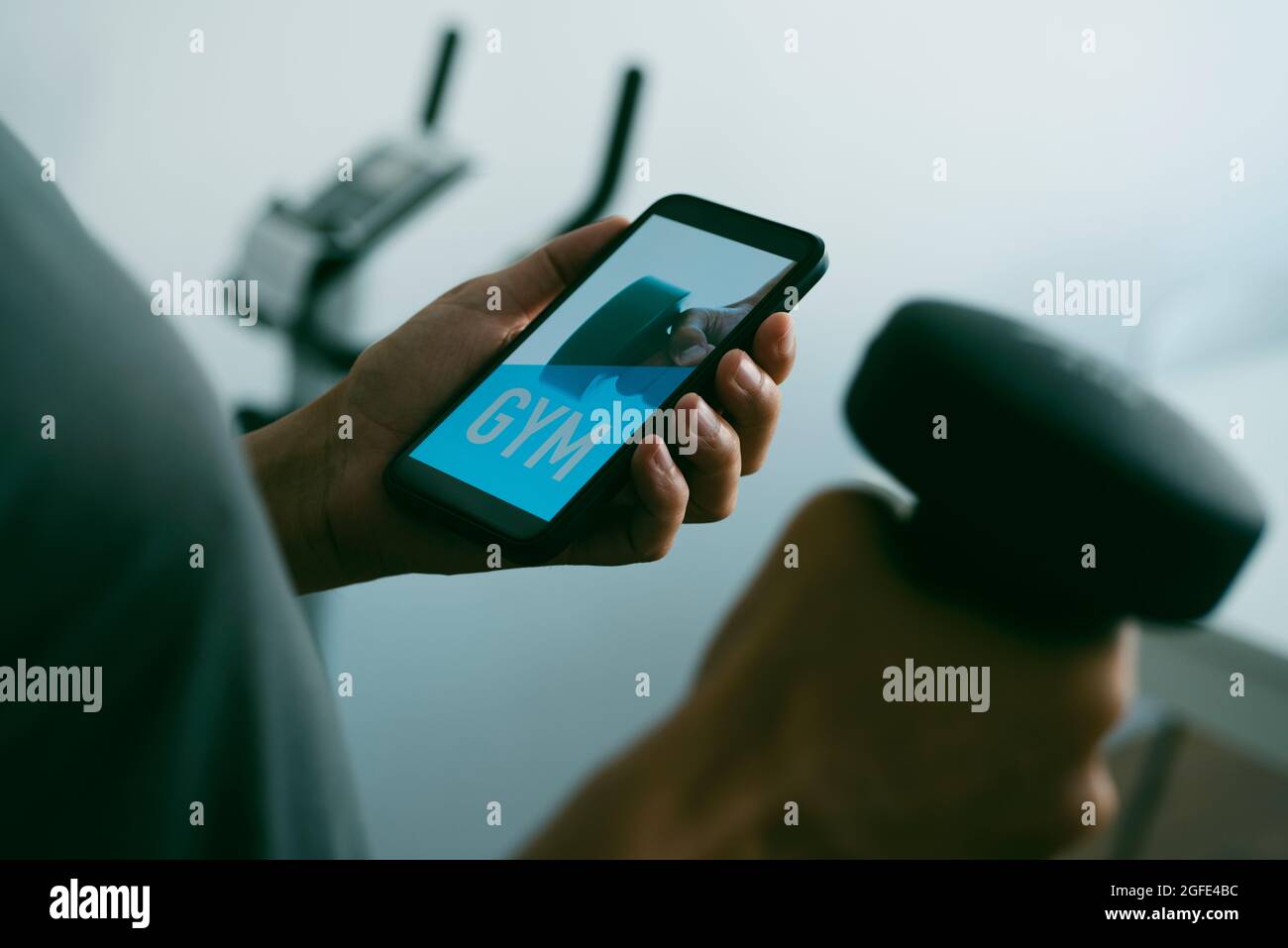 primo piano di un giovane uomo caucasico che solleva un manubano nel soggiorno di casa mentre guarda al suo smartphone, che legge palestra nel suo schermo, Foto Stock