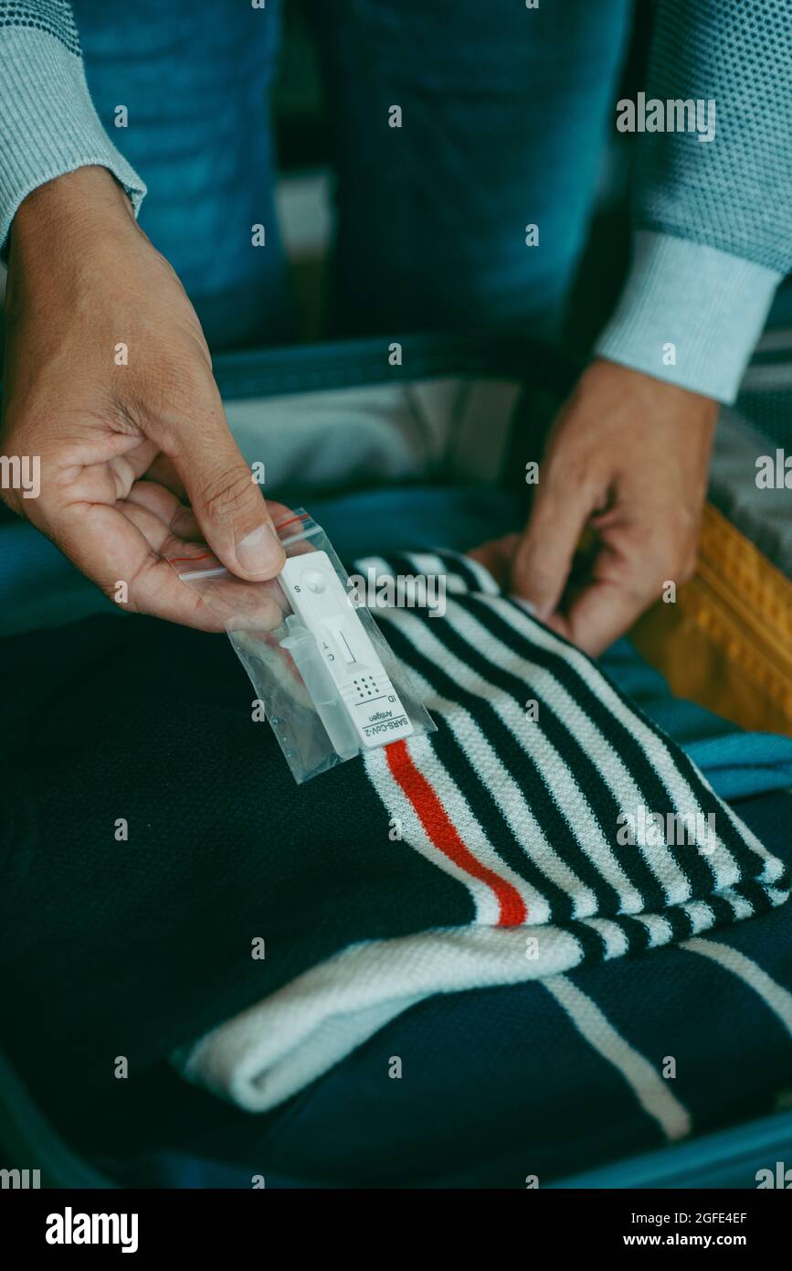 primo piano di un giovane uomo caucasico che impacchette un covid-19 kit di test diagnostico antigenico nella sua valigia, confezionato con diversi vestiti, sul letto Foto Stock