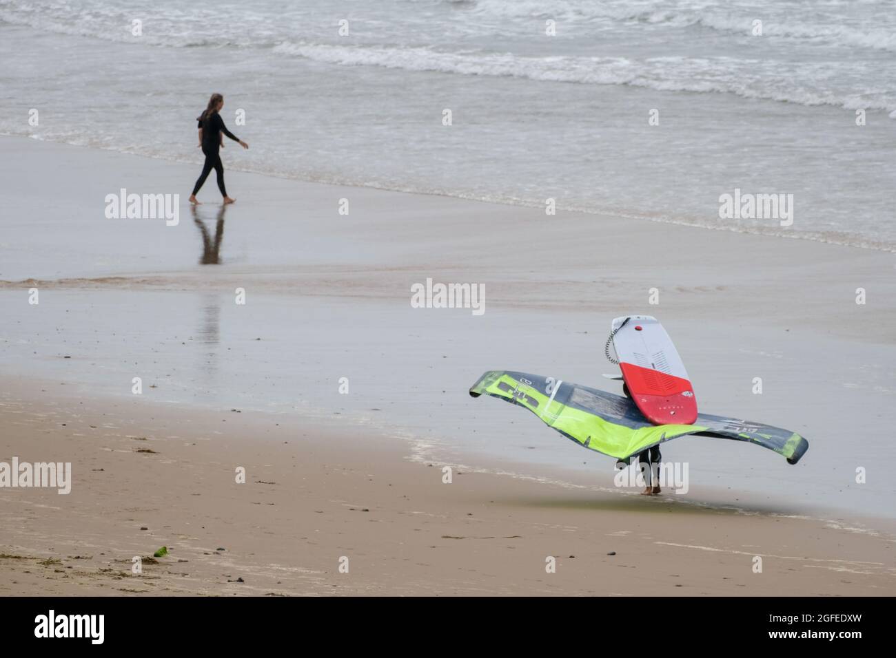 Una persona che trasporta una scheda di foglio; quella è una tavola da surf di aliscafo usata normalmente in congiunzione con un aquilone per propulsione, fa il suo senso lungo la spiaggia di Llangennith, Gower, Swansea, Regno Unito, verso il mare. Foto Stock