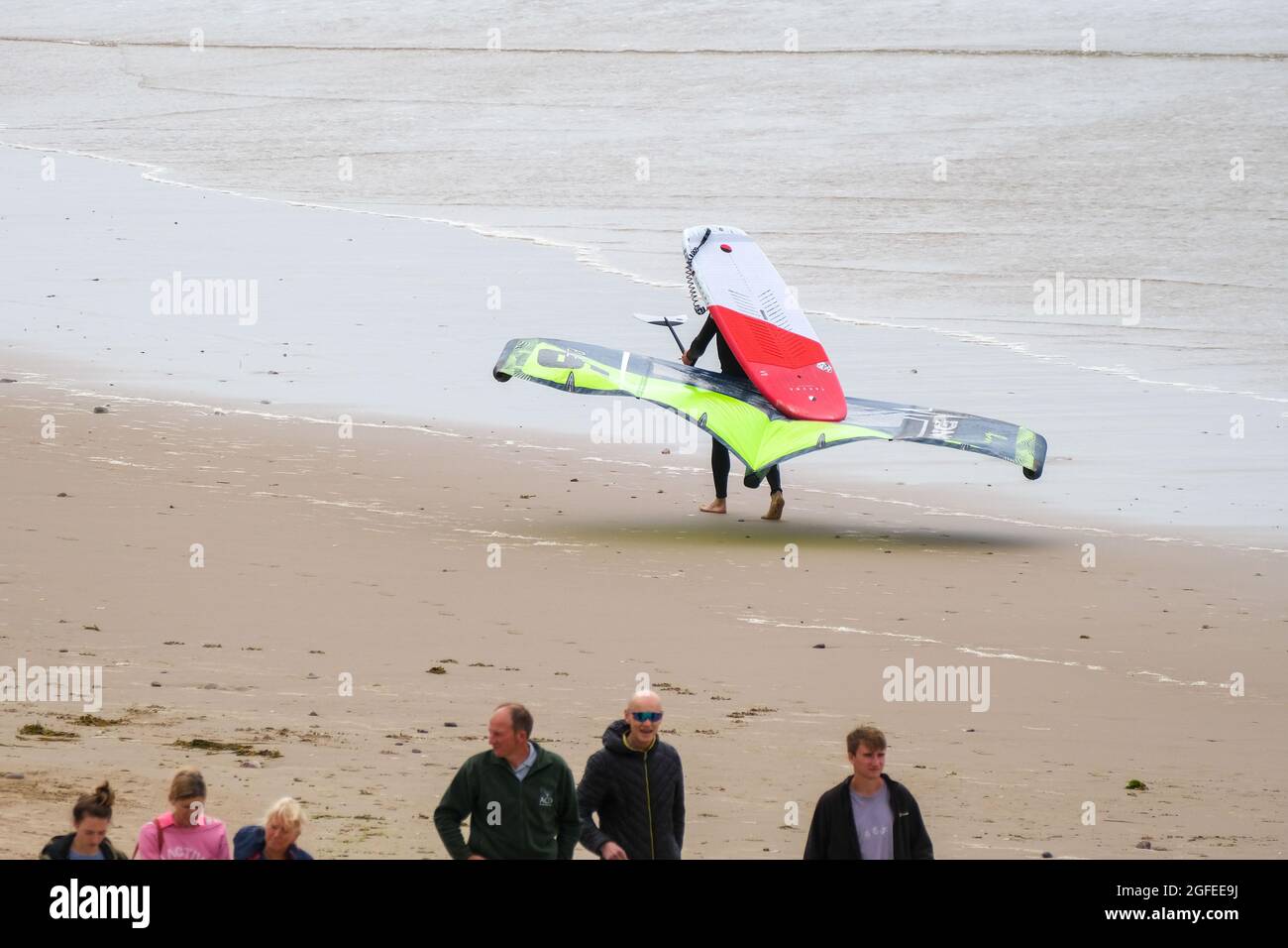 Una persona che trasporta una scheda di foglio; quella è una tavola da surf di aliscafo usata normalmente in congiunzione con un aquilone per propulsione, fa il suo senso lungo la spiaggia di Llangennith, Gower, Swansea, Regno Unito, verso il mare. Foto Stock