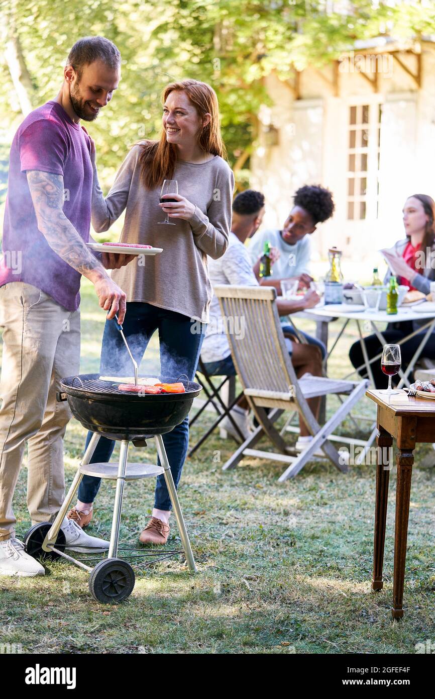 Giovane uomo che prepara il cibo in barbecue grill con i suoi amici seduti a tavola sullo sfondo Foto Stock