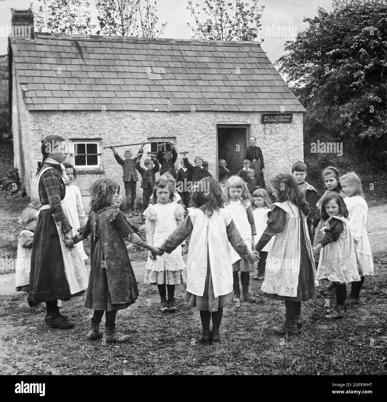 Bambini in gioco durante il 20 ° secolo alla Scuola Nazionale Ballidiana, Ballybay nella contea di Monaghan, Irlanda. Le ragazze che giocano a “Green Grow the rushes, o” mentre i ragazzi fanno esercizi ginnici sorvegliati dal loro insegnante. Foto Stock