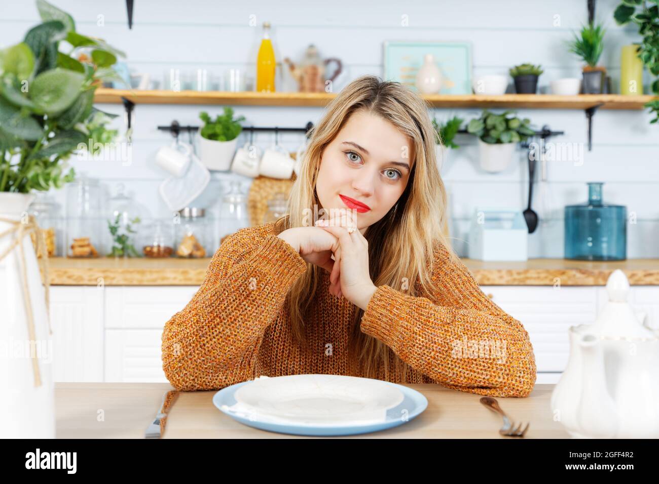 Giovane donna seduta al tavolo da cucina di fronte ad un piatto vuoto. Sfondo sfocato. Foto Stock