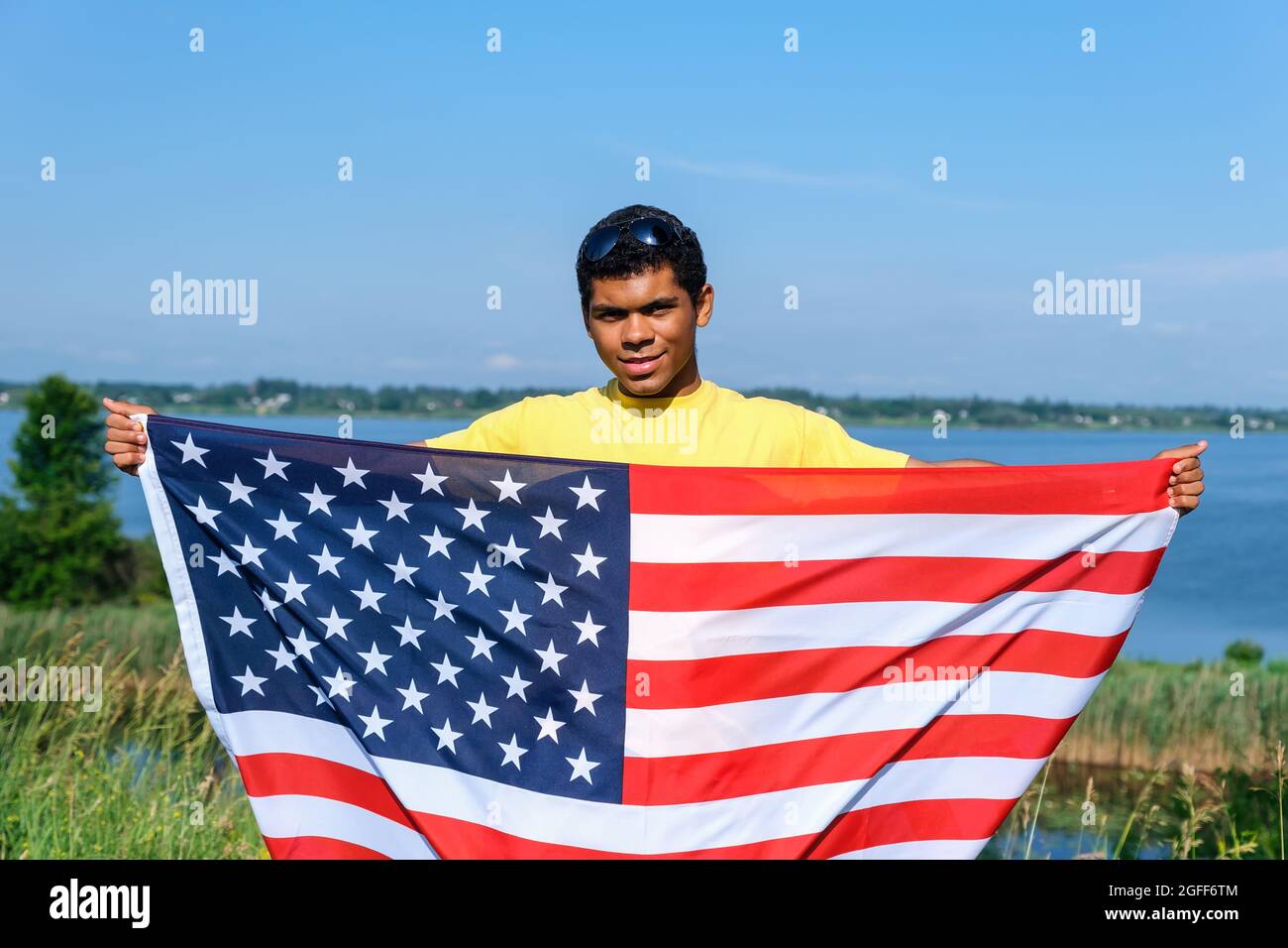 Giovane afroamericano con bandiera USA in piedi vicino al fiume in estate soleggiata Foto Stock