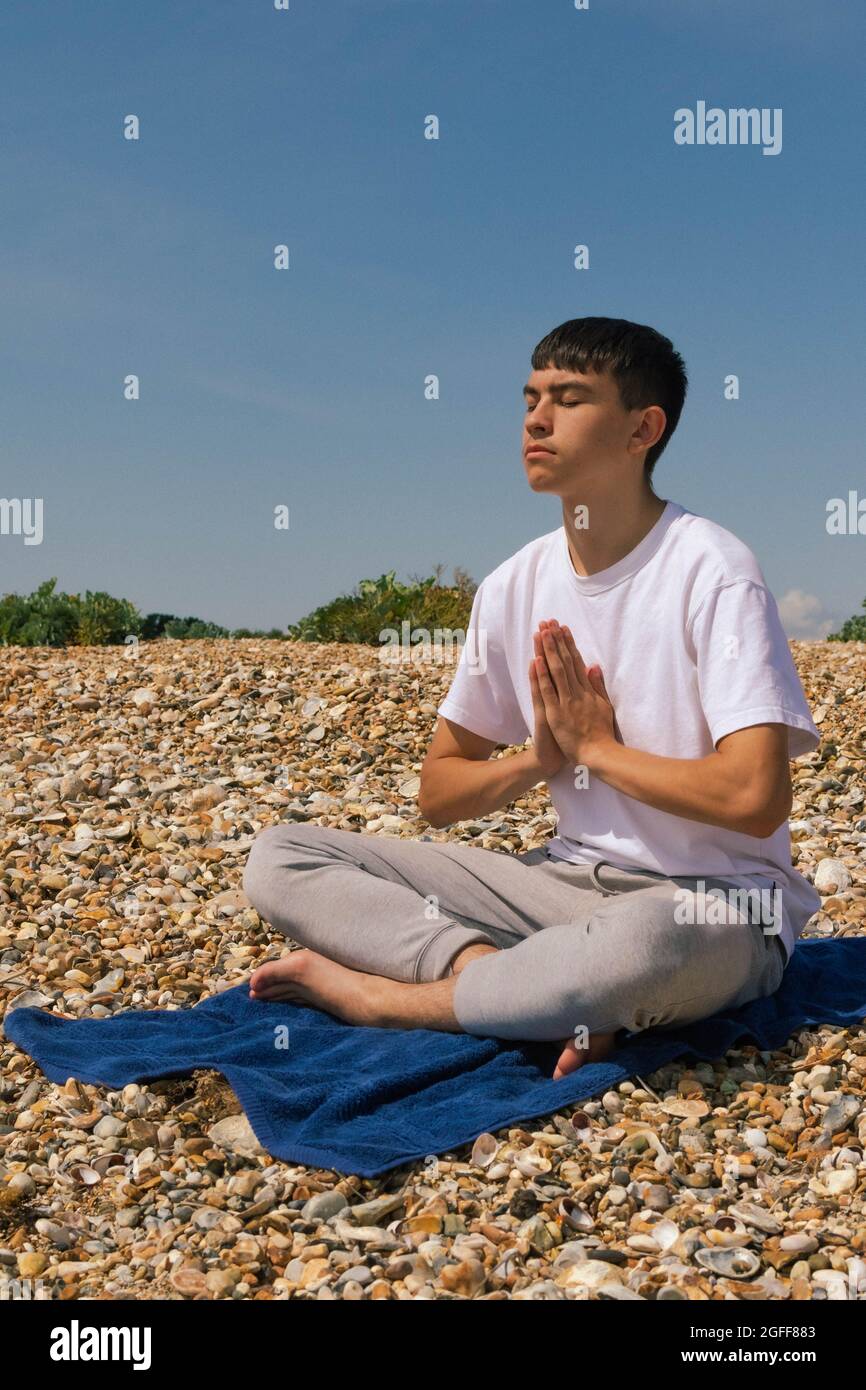 Un adolescente caucasico meditando su una spiaggia di pietra con le mani in una posizione di preghiera Foto Stock