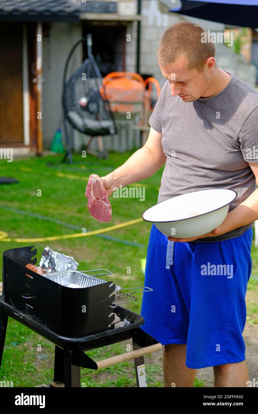Ritratto di un giovane che grigliava carne in Polonia. Vista a metà lunghezza di un giovane caucasico che mette una bistecca sul grill esterno Foto Stock