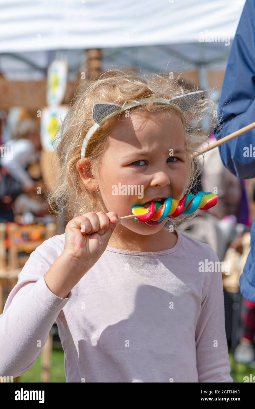 Ragazza che mangia caramelle colorate. Una ragazza di cinque anni in vestiti rosa sta mangiando una caramella colorata su un bastone. Giornata estiva soleggiata Foto Stock