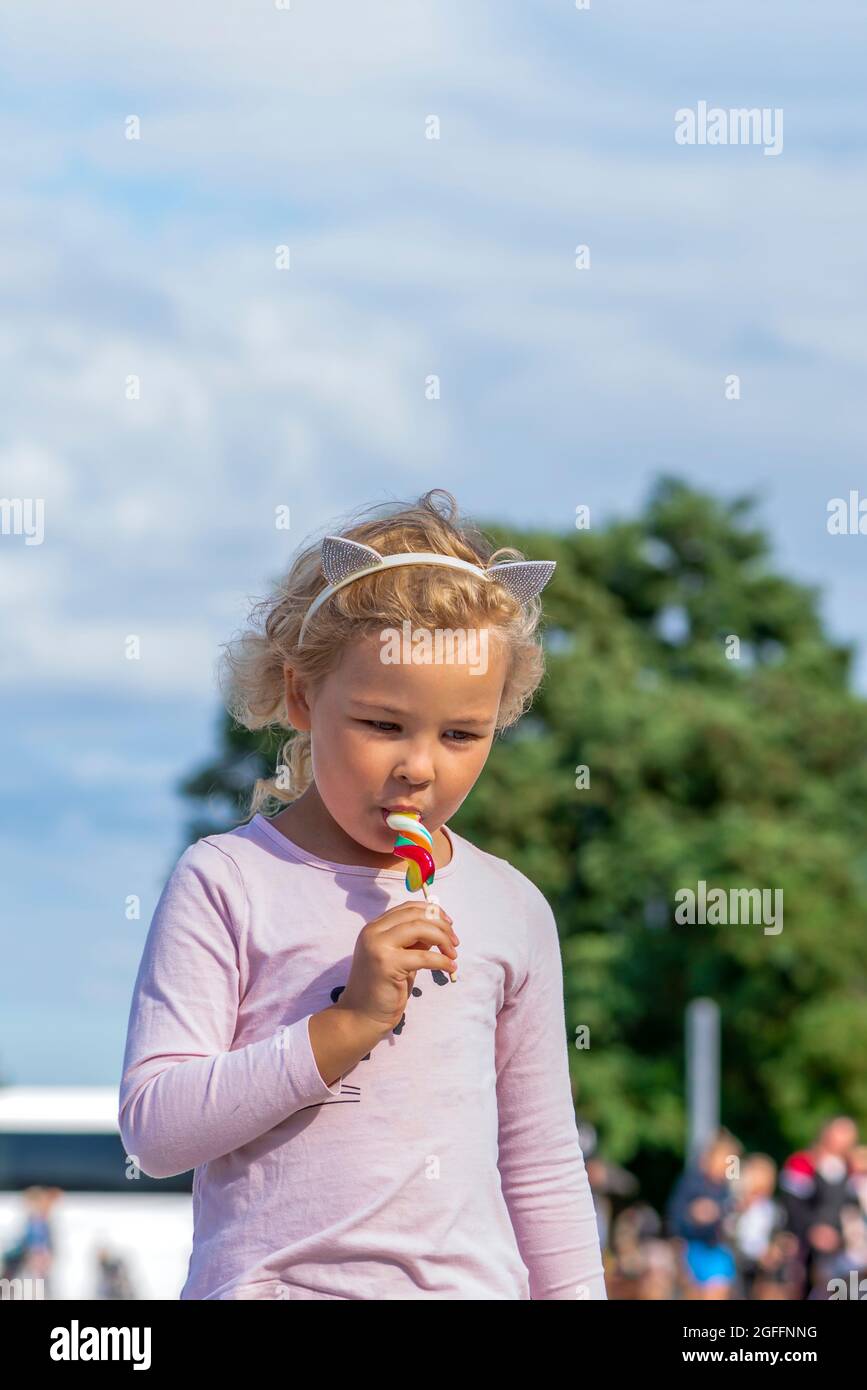 Ragazza che mangia caramelle colorate. Una ragazza di cinque anni in vestiti rosa sta mangiando una caramella colorata su un bastone. Giornata estiva soleggiata Foto Stock