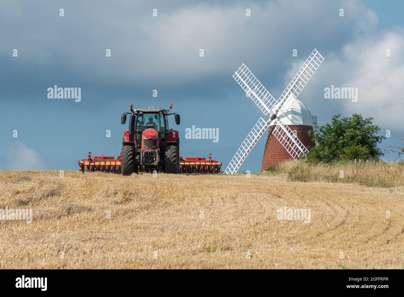 Hornaker Windmill nel South Downs National Park nel West Sussex, Inghilterra, Regno Unito, con un trattore rosso che lavora nel campo di mais, in una giornata estiva soleggiata Foto Stock