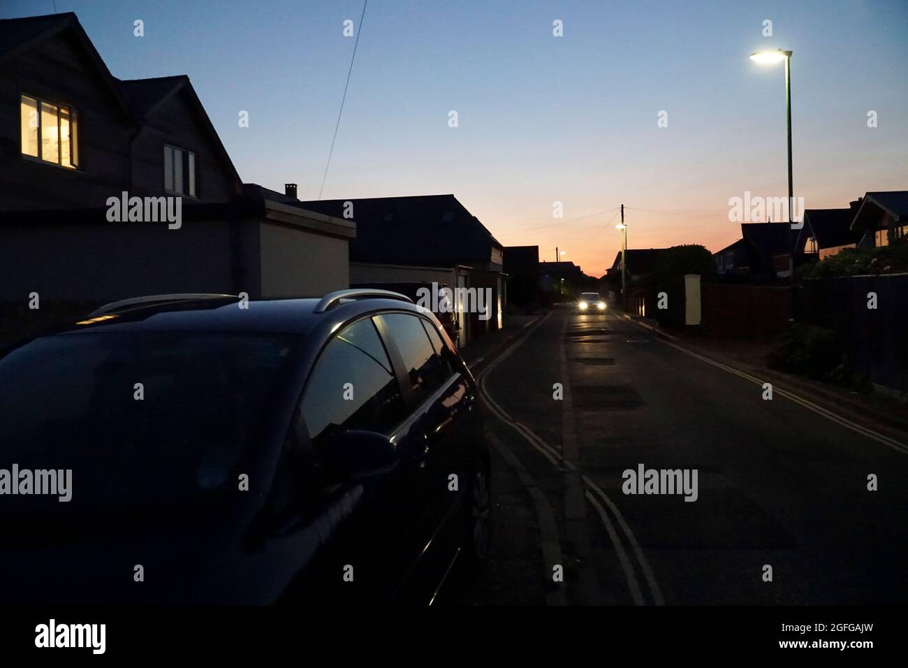 East Wittering, UK, 25 agosto 2021: Un cielo blu e rosa dà un'ultima luce, riflessa da un'auto parcheggiata, come finestre di casa, luci di strada e ca Foto Stock
