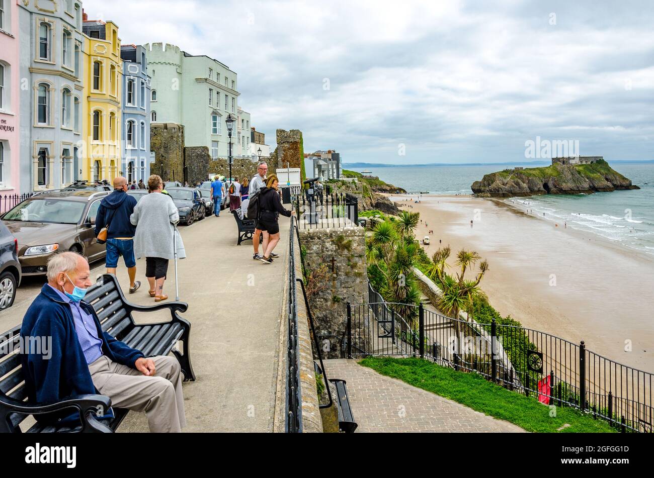 Un uomo si siede sulla panchina dell'Esplanade a Tenby, leggendo con la South Beach e l'Isola di Santa Caterina in lontananza. Foto Stock