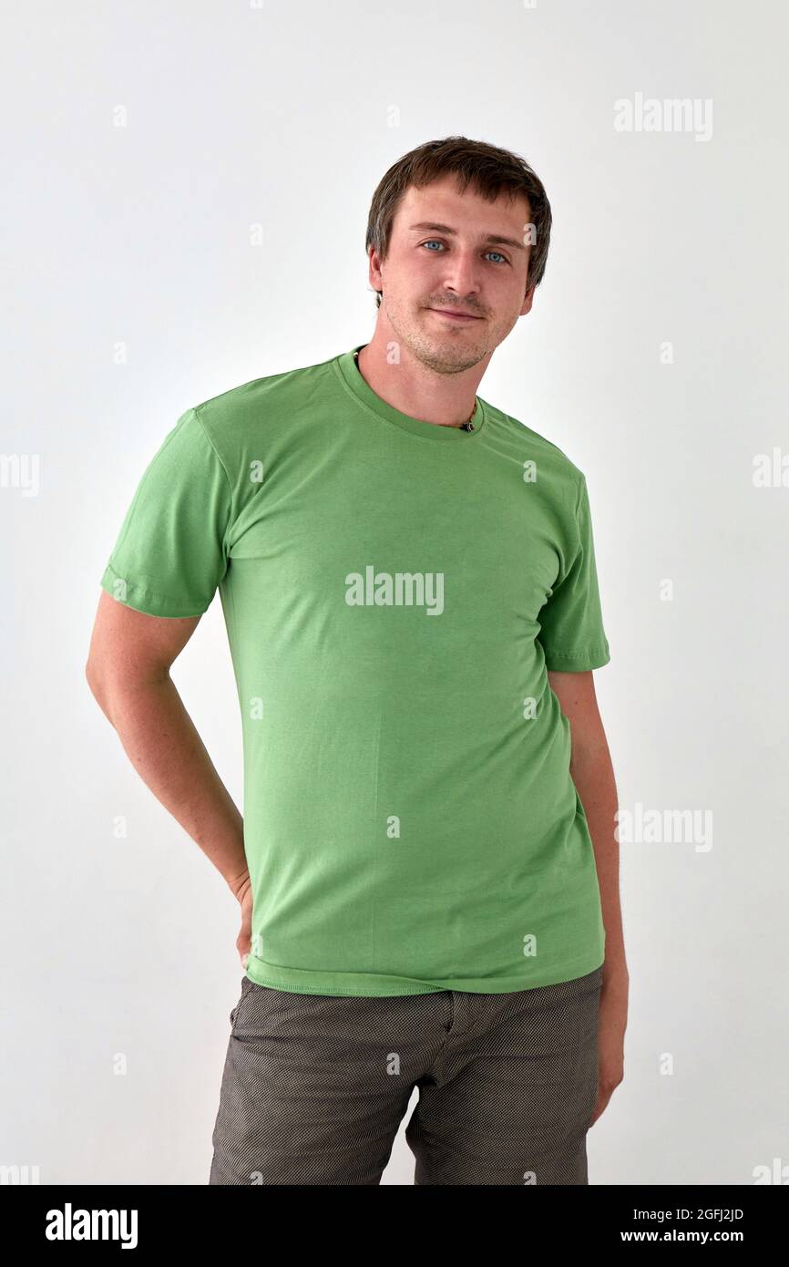 Positivo giovane maschio in jeans con mano dietro e t shirt verde in piedi su sfondo bianco e guardando la macchina fotografica Foto Stock