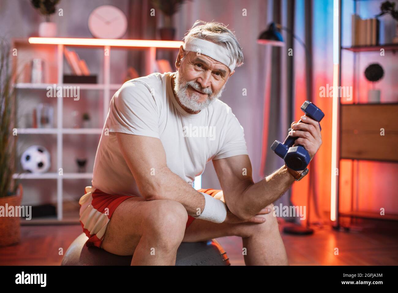 Uomo anziano sorridente di 70 anni in abbigliamento sportivo e fasciatesta  seduto su palla svizzera e braccia da training con manubri. Pensionato  caucasico che fa un allenamento regolare a casa. Stili di