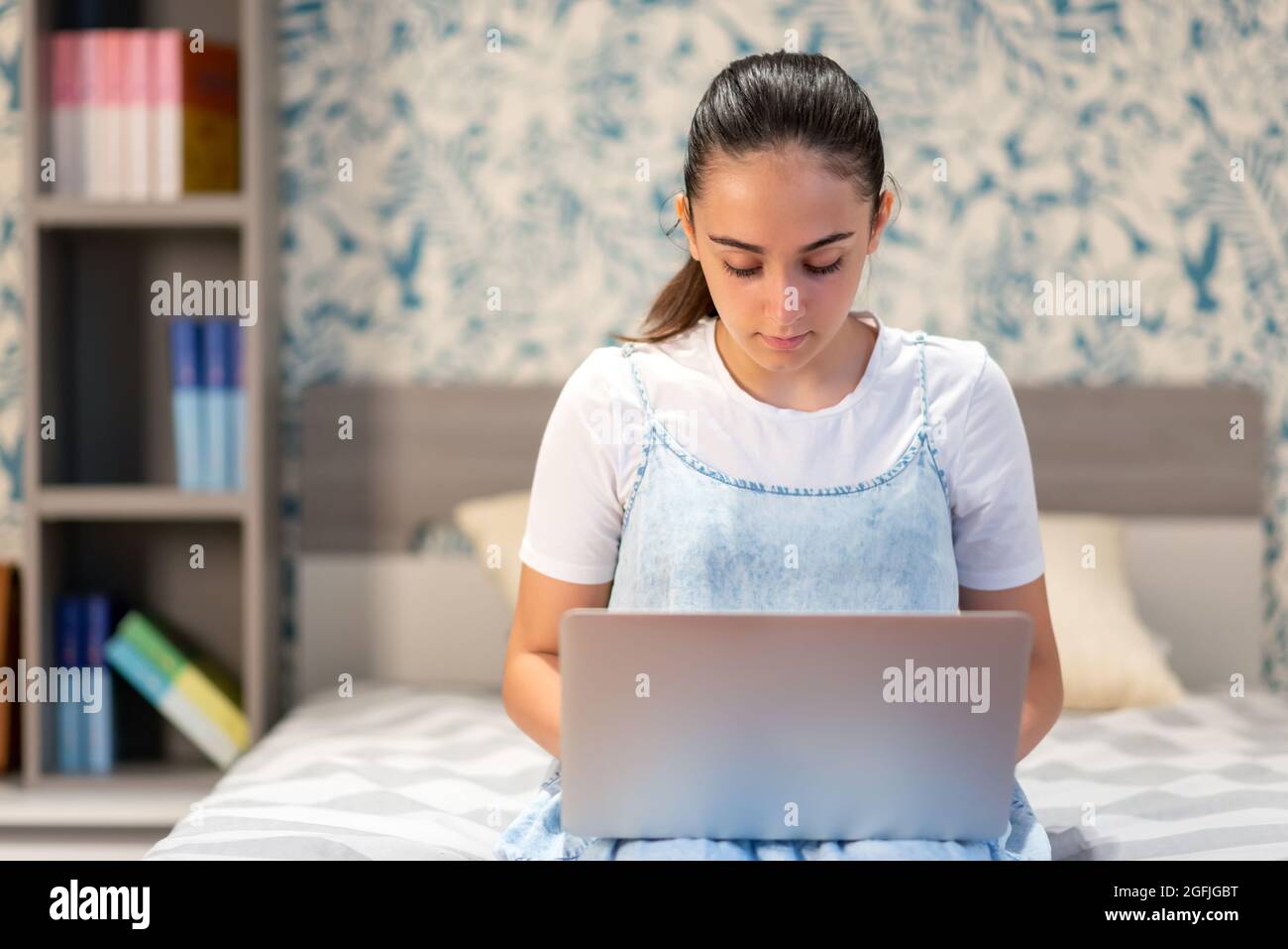 Ragazza seria teen seduta a letto e utilizzando un computer portatile mentre comunica online durante il tempo libero a casa Foto Stock