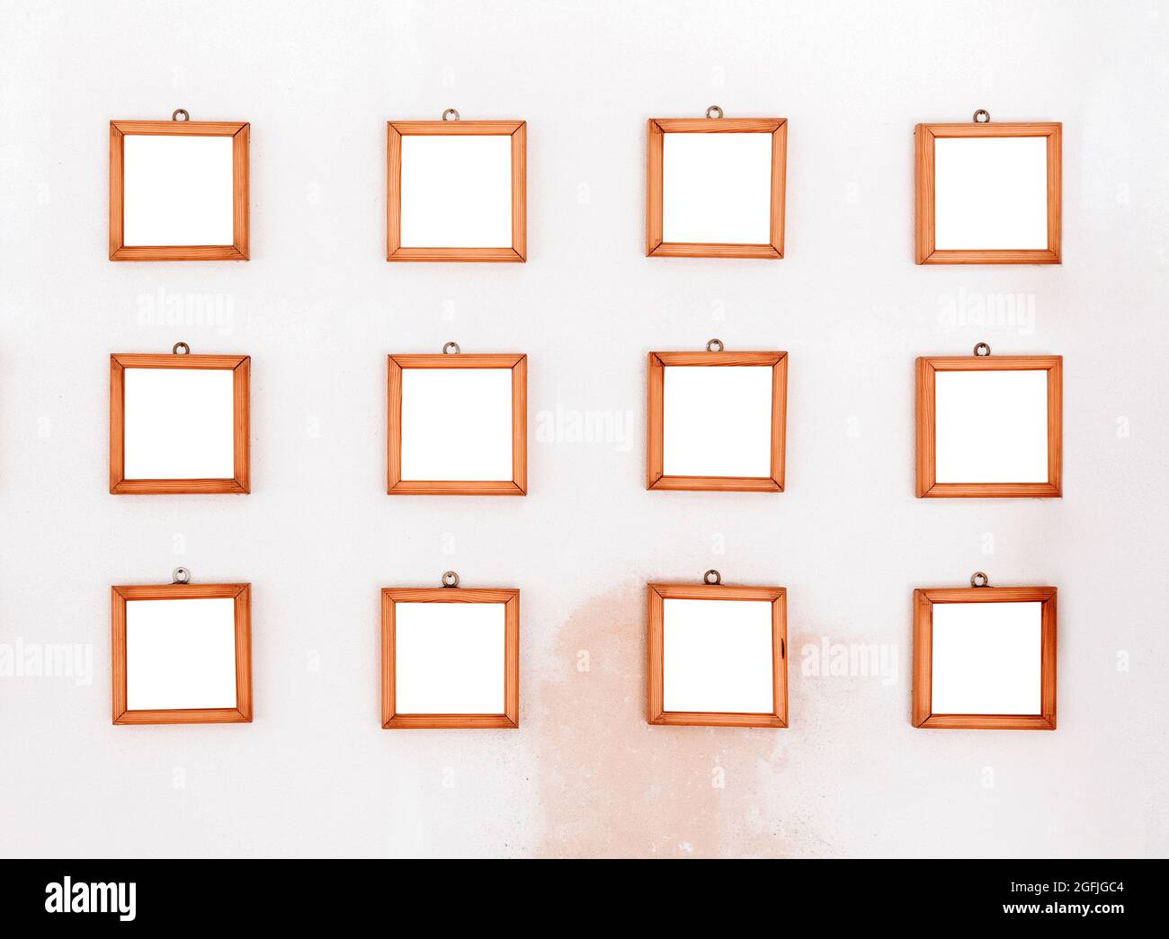Dodici piccole cornici di legno quadrate semplici e vuote esposte su una parete bianca con macchia rossastra scolorita sotto in tre file pronte per uno spettacolo o una mostra Foto Stock