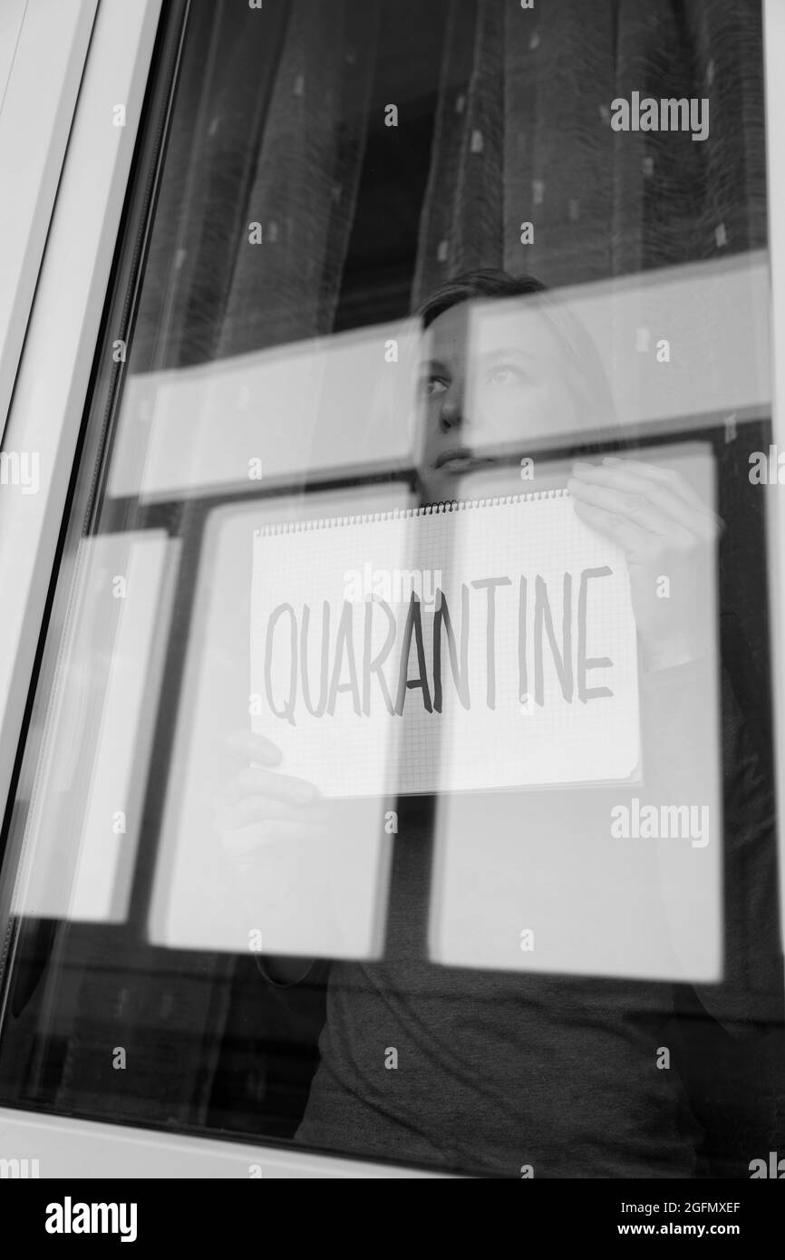 Donna che guarda attraverso la finestra e che tiene un cartello scritto a mano che dice 'Quarantine'. Verticale in bianco e nero. Foto Stock