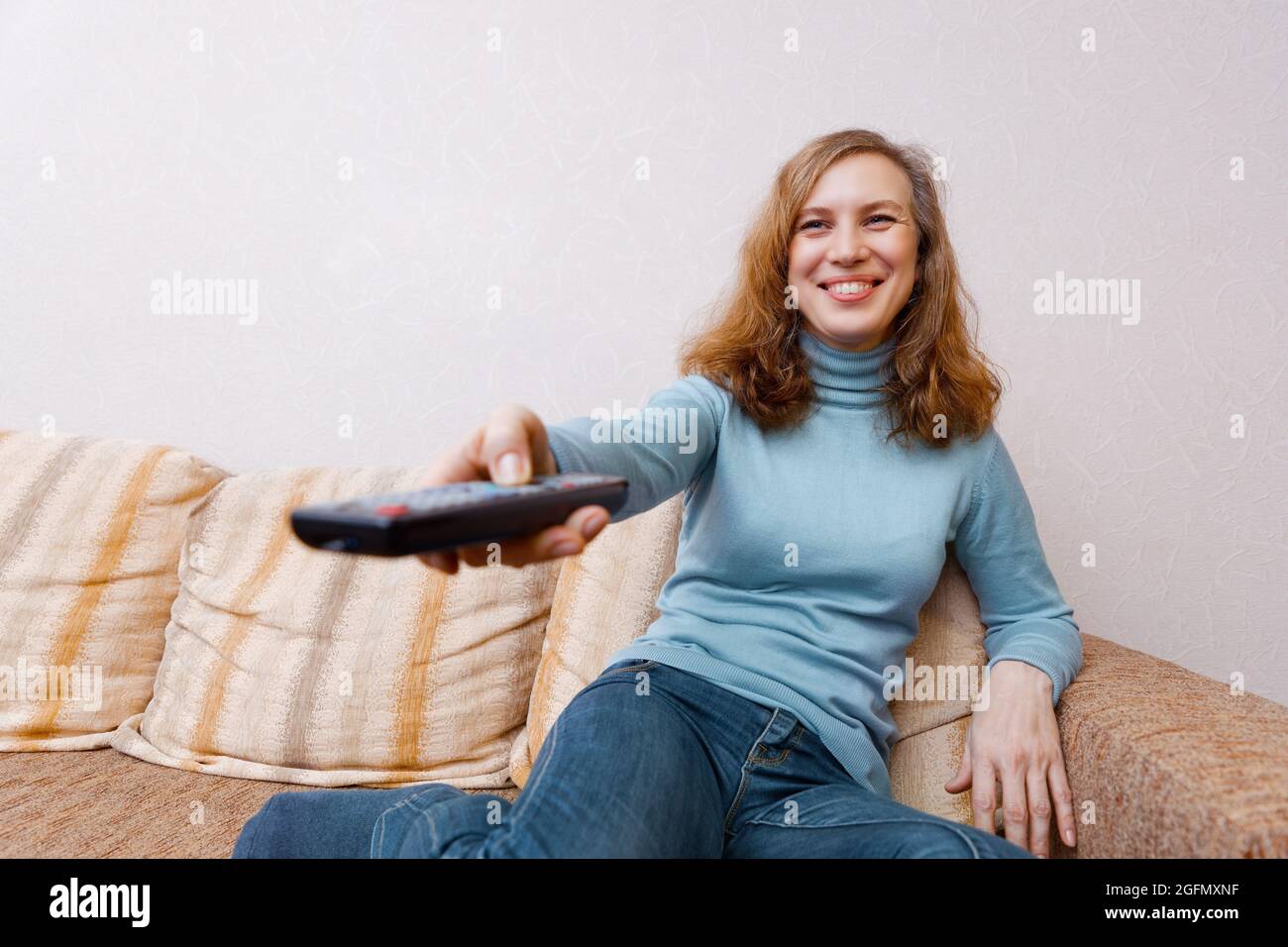 Donna sorridente positiva seduta sul divano a casa guardare la tv con telecomando. Messa a fuoco superficiale. Foto Stock