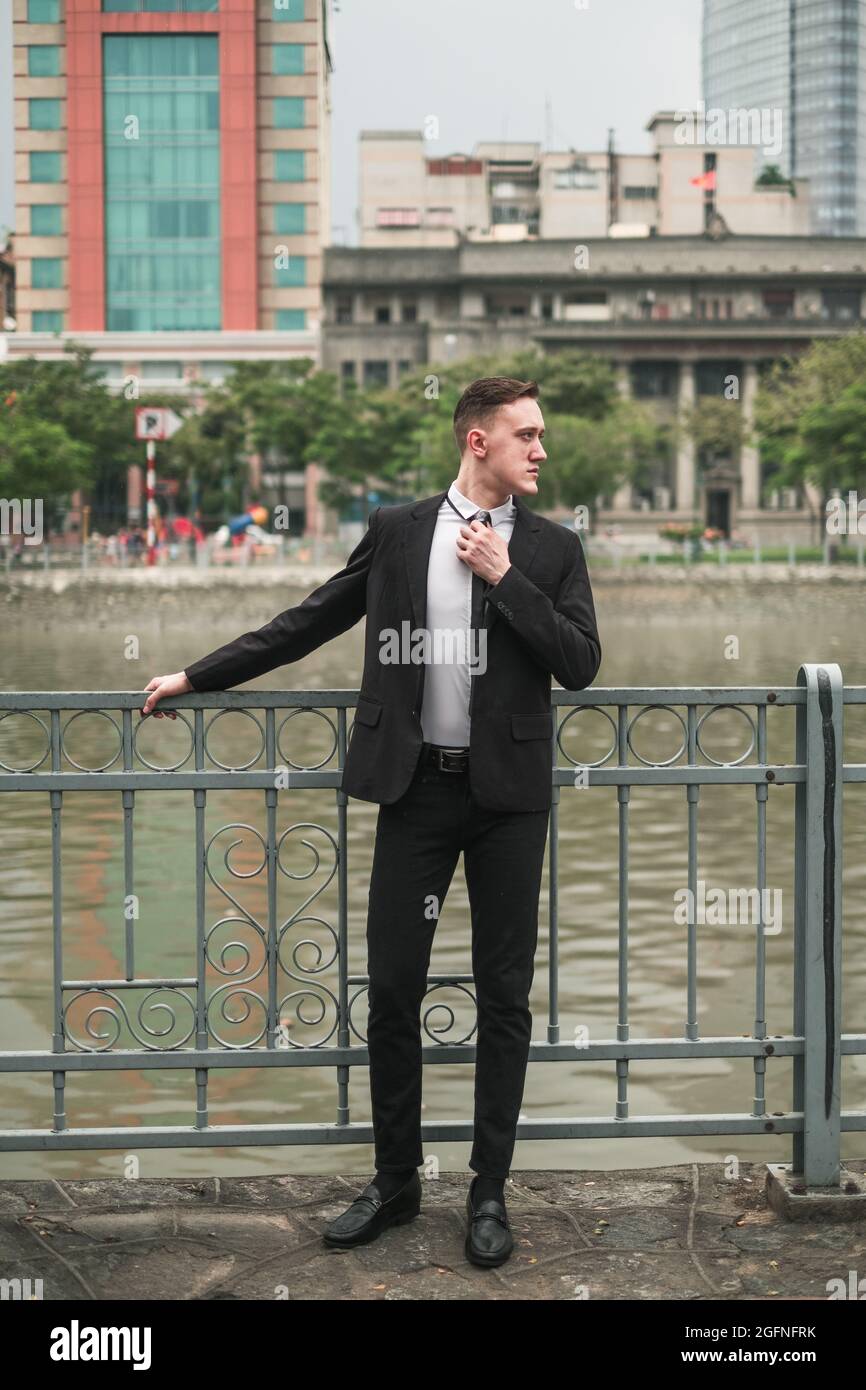 Bel giovane uomo in abito nero formale in piedi vicino al fiume nella grande città. Raddrizza la cravatta. Grattacieli sullo sfondo Foto Stock