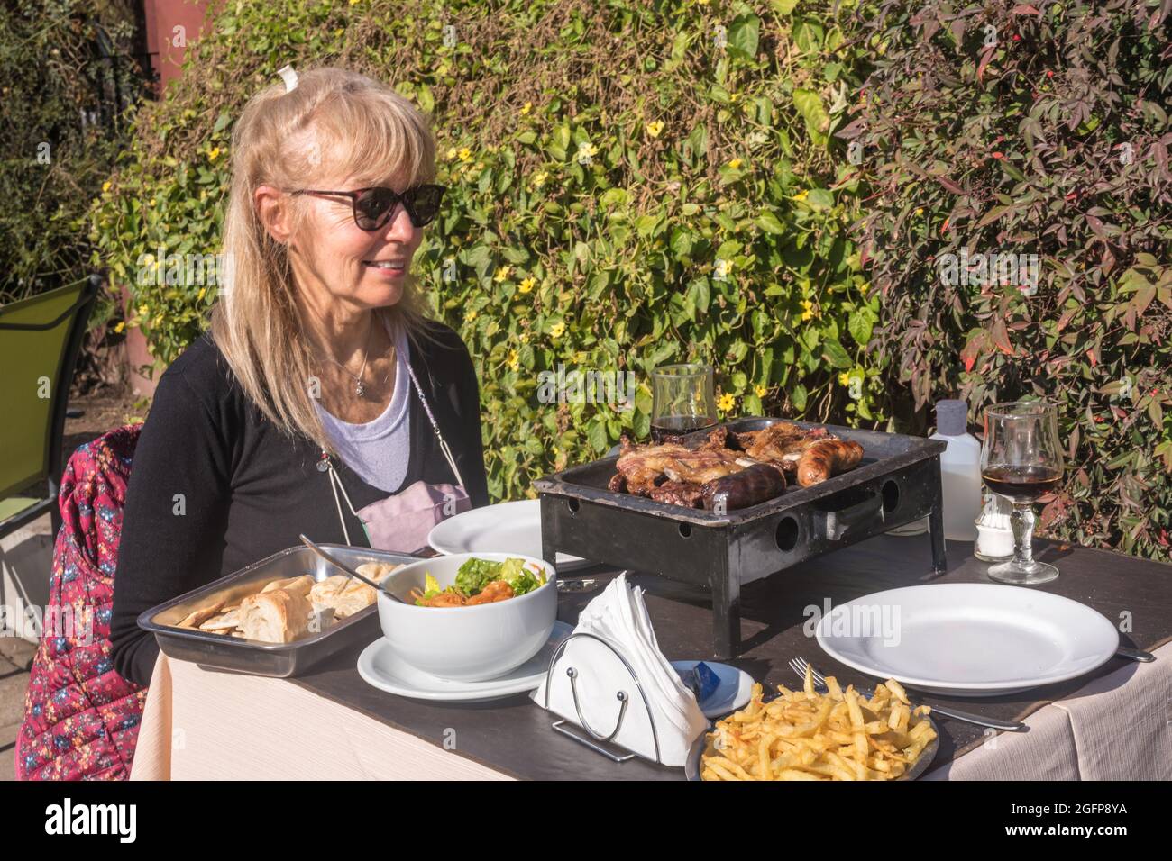 Donna adulta seduta al tavolo sorridente guardando barbecue argentino, insalata e patatine fritte. Foto Stock