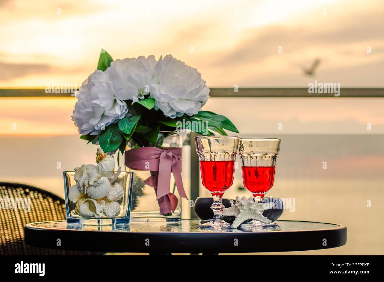 due bicchieri di vino sullo sfondo del tramonto sul mare, su un tavolo sul balcone, conchiglie e fiori Foto Stock