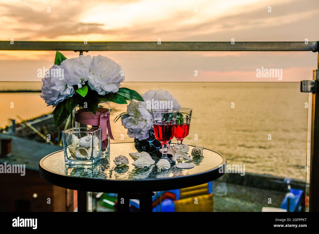 due bicchieri di vino sullo sfondo del tramonto sul mare, su un tavolo sul balcone, conchiglie e fiori Foto Stock