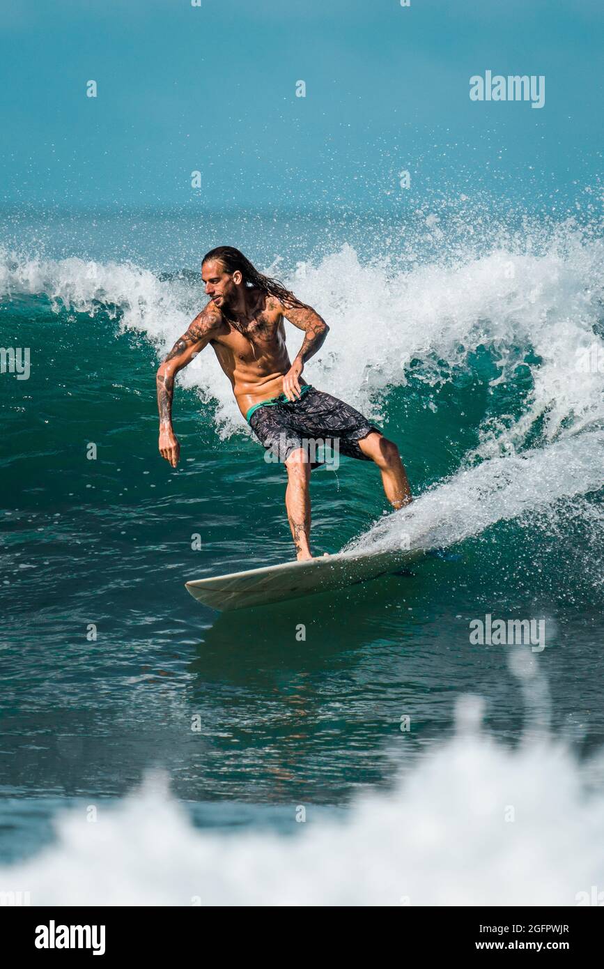 Una bella foto di un uomo dai capelli lunghi con tatuaggi che cavalcano casualmente un'onda sulla costa pacifica del Costa Rica Foto Stock