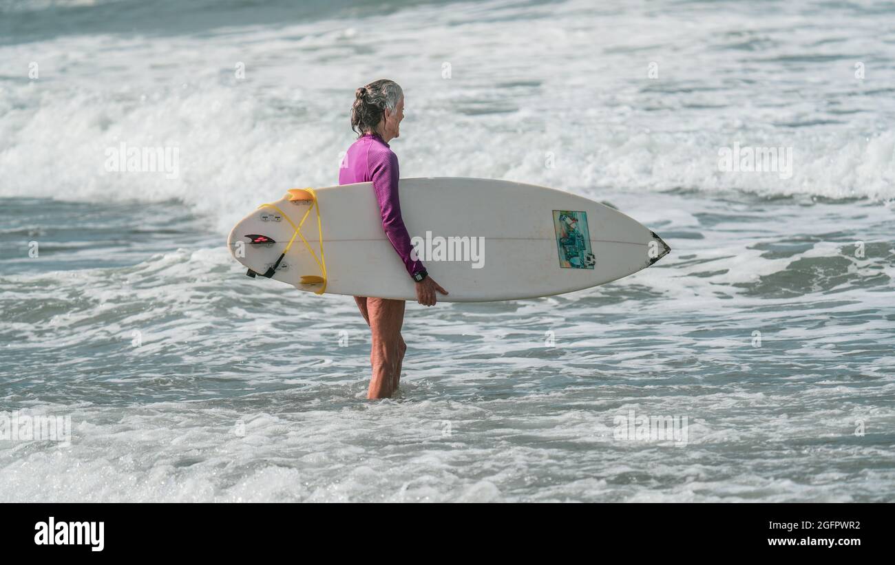 Una vecchia donna attiva con capelli grigi sta tenendo una tavola da surf e sorridendo mentre si prepara a fare surf sulla costa pacifica del Costa Rica Foto Stock