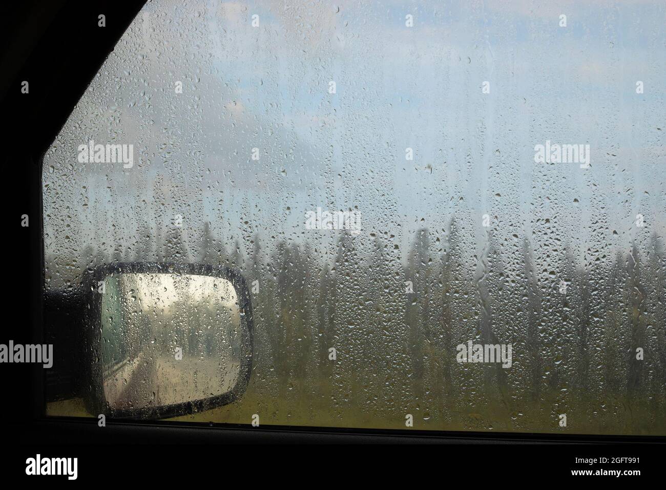 Gocce di pioggia sul finestrino di un veicolo, guardando verso lo specchietto retrovisore, la foresta e il cielo nuvoloso durante un forte temporale Foto Stock
