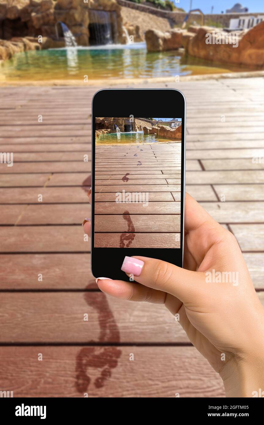 Immagine ravvicinata del telefono cellulare con le mani femminili e la modalità fotocamera sullo schermo. Immagine ritagliata delle impronte sul pavimento di legno dietro di esso Foto Stock