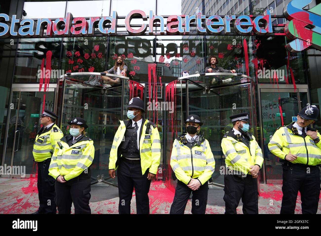 I dimostranti si siedono sopra le porte dell'edificio Standard Chartered, che è stato scolorito, mentre gli ufficiali di polizia bloccano l'accesso mentre una protesta della ribellione dell'estinzione attraversa la città di Londra. Data foto: Venerdì 27 agosto 2021. Foto Stock