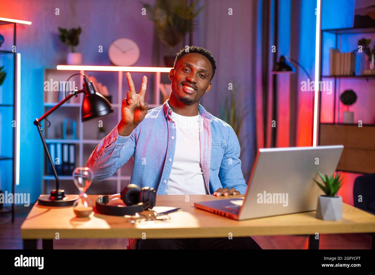 Bell'uomo africano seduto al tavolo con un moderno computer portatile e gesturante segno di pace con le dita. Ragazzo felice che si gode di lavoro a distanza a casa. Concetto di freelance e tecnologia. Foto Stock