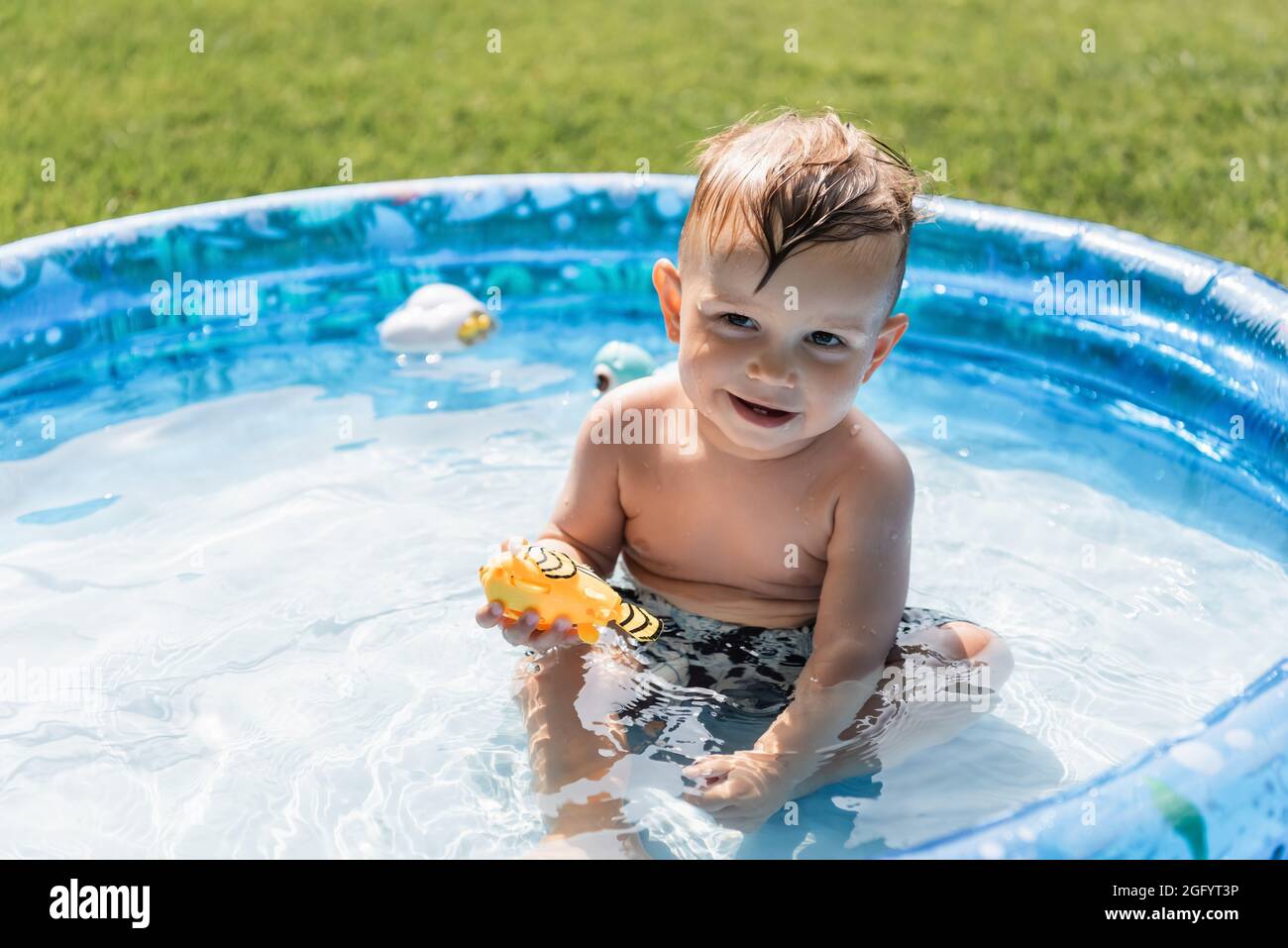 il ragazzo positivo del toddler che siede nella piscina gonfiabile e che tiene il giocattolo di gomma Foto Stock