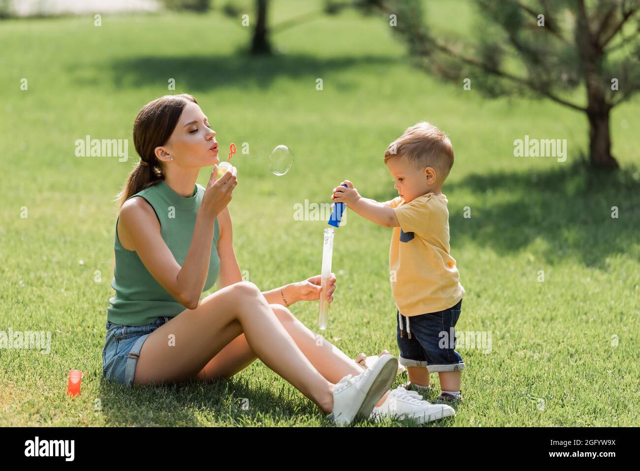 madre seduta su erba e soffiare bolla di sapone vicino bambino ragazzo Foto Stock