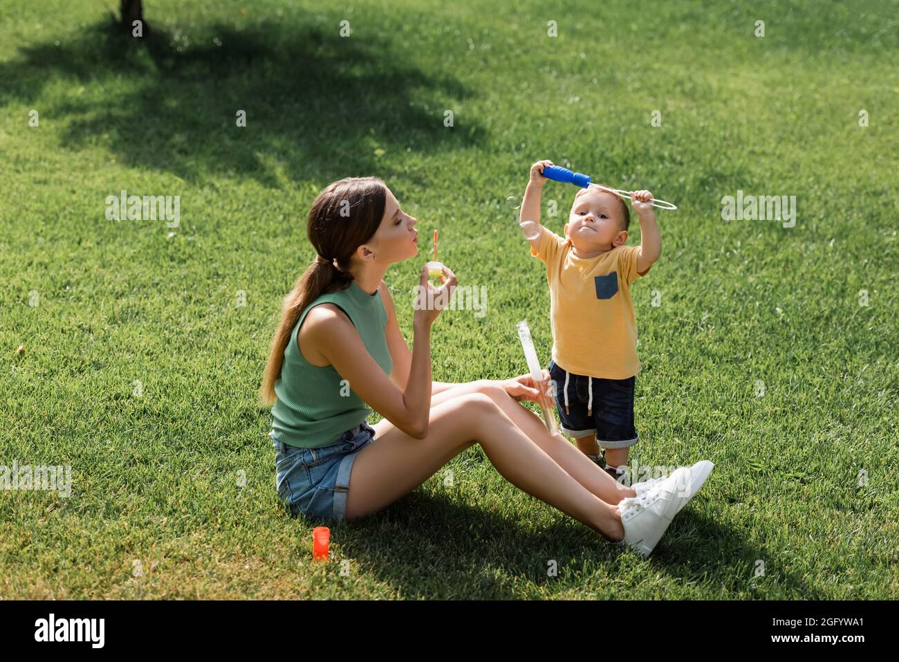 madre giovane seduta su erba e bolle di sapone soffiato vicino figlio del toddler Foto Stock