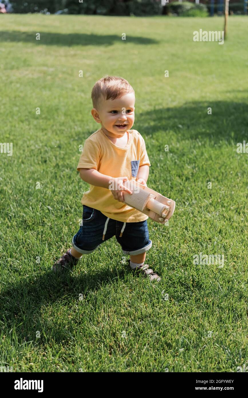 ragazzo allegro con auto giocattolo in legno mentre cammina sull'erba Foto Stock