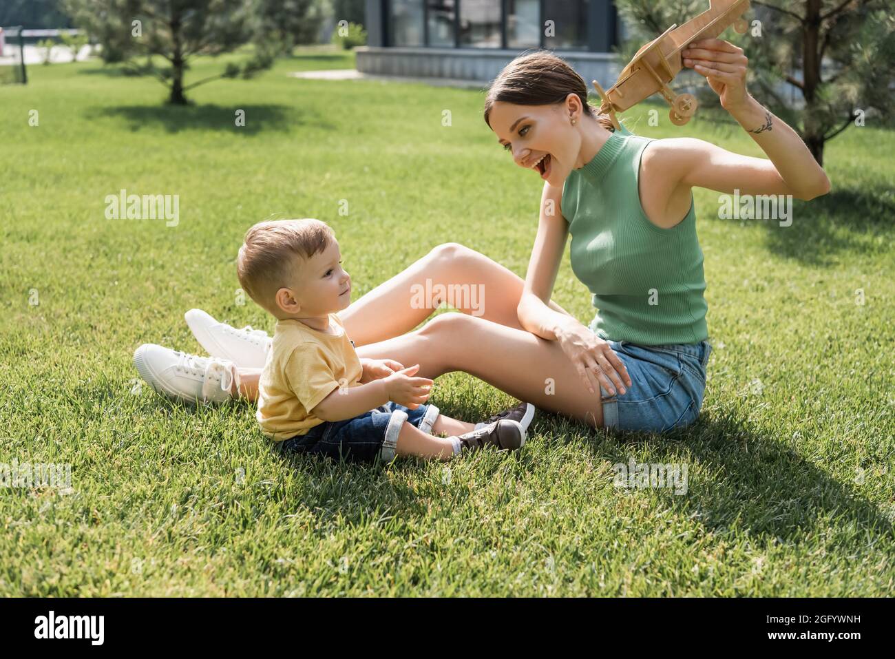 felice madre giovane tenendo giocattolo biplanare e guardando il figlio allegro toddler su erba Foto Stock