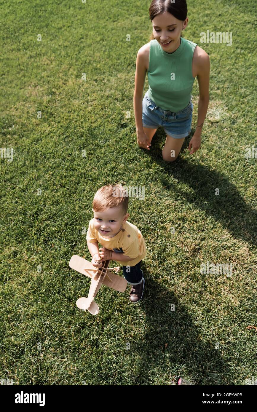 vista dall'alto della giovane madre felice che guarda il figlio allegro del bambino con biplano giocattolo e in piedi sull'erba Foto Stock