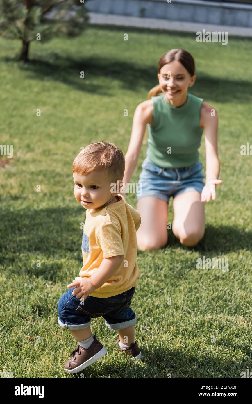 mamma allegra e sfocata con le mani distese che si siedono sull'erba e raggiungono il figlio del bambino fuori Foto Stock