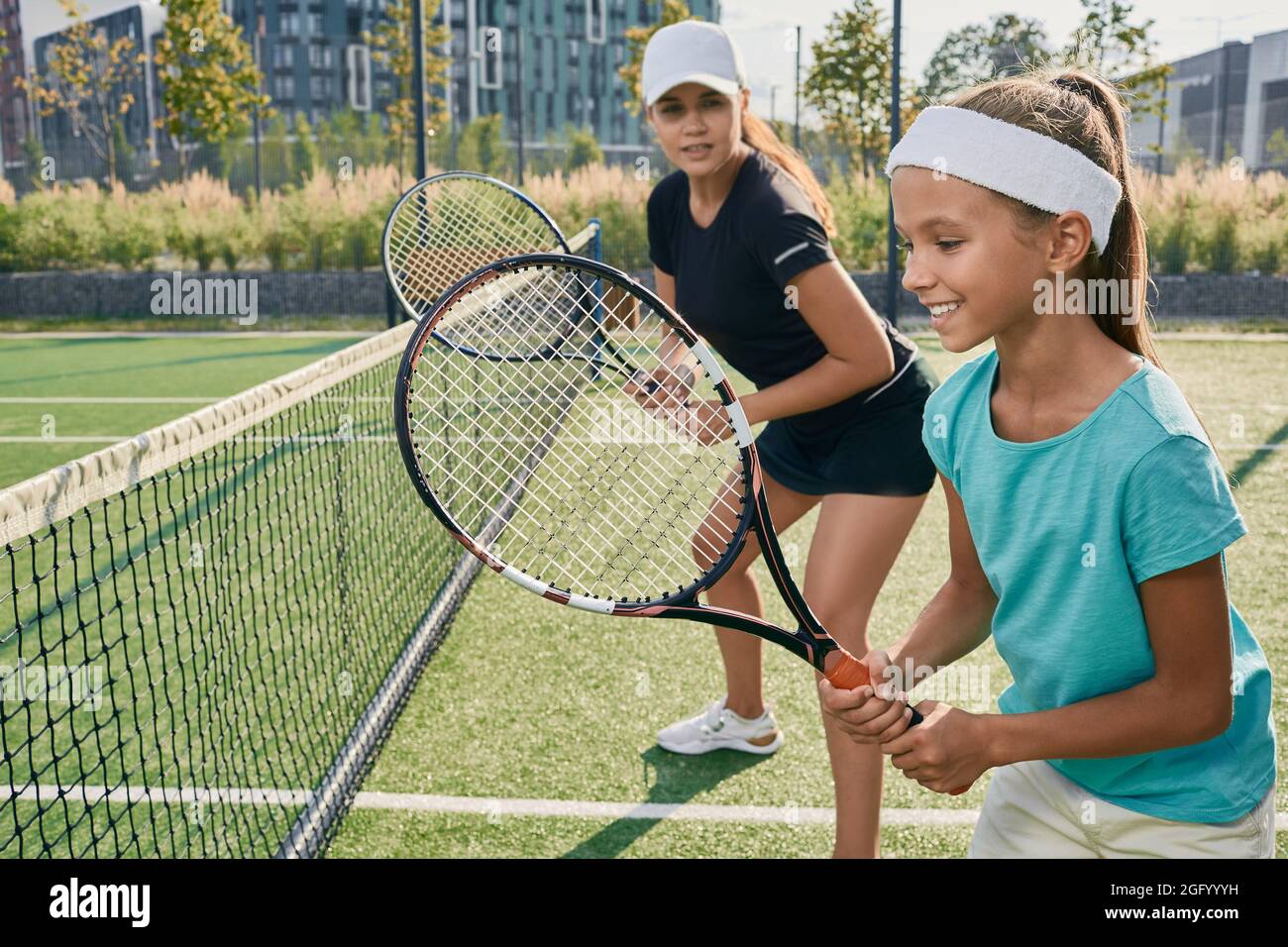 Ragazza piccola positiva che tiene un racket mentre impara a giocare a tennis con il suo allenatore femminile sul campo all'aperto. Istruttore individuale a un bambino per i tenni Foto Stock