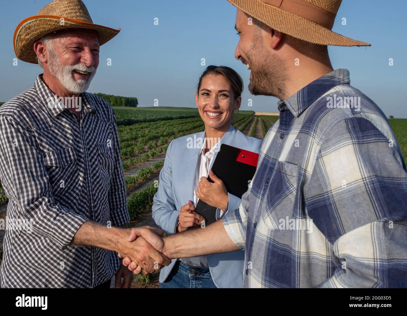 Tre persone in piedi sul campo che fanno affari raggiungere un accordo. Giovane rappresentante di vendita di assicurazione femminile che tiene appunti. Due agricoltori che scuotono h Foto Stock