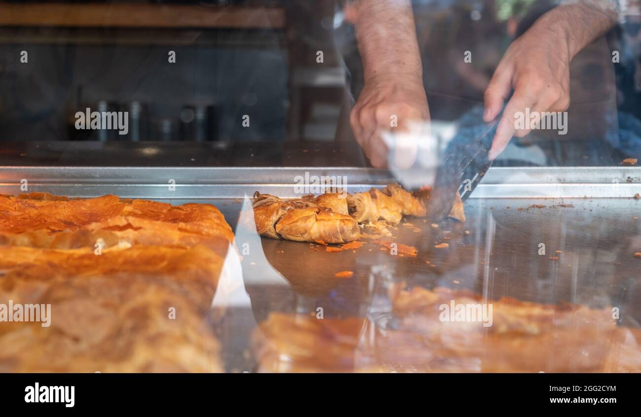Bougatsa in una vetrina, Male mani tagliando pasticceria con carne macinata a pezzi. Colazione tradizionale con ripieno dolce o salato tra strati di Foto Stock