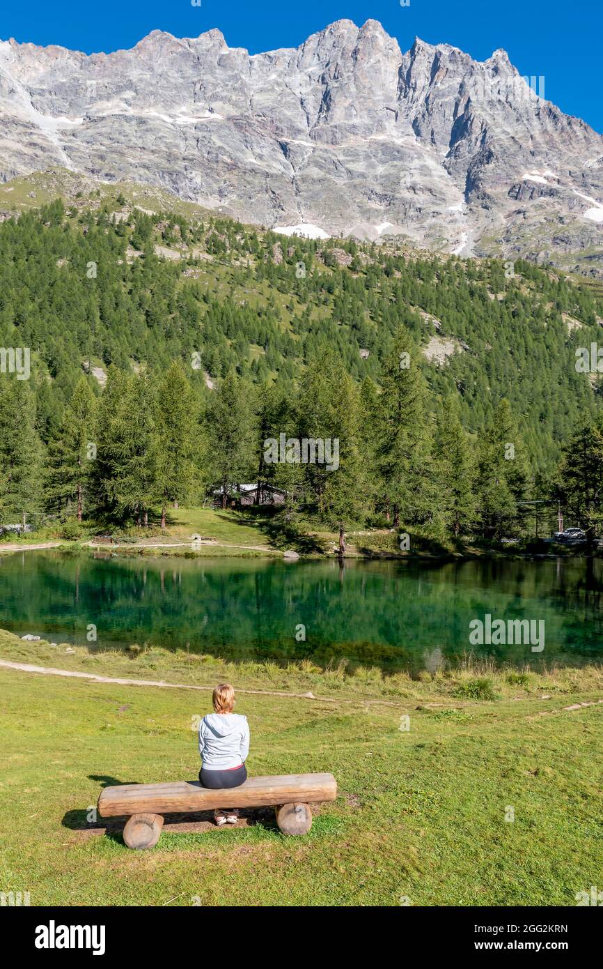 Una donna bionda seduta su una panchina di legno ammira il panorama del Lago Blu in cui si riflettono le montagne circostanti, Valle d'Aosta, Italia Foto Stock