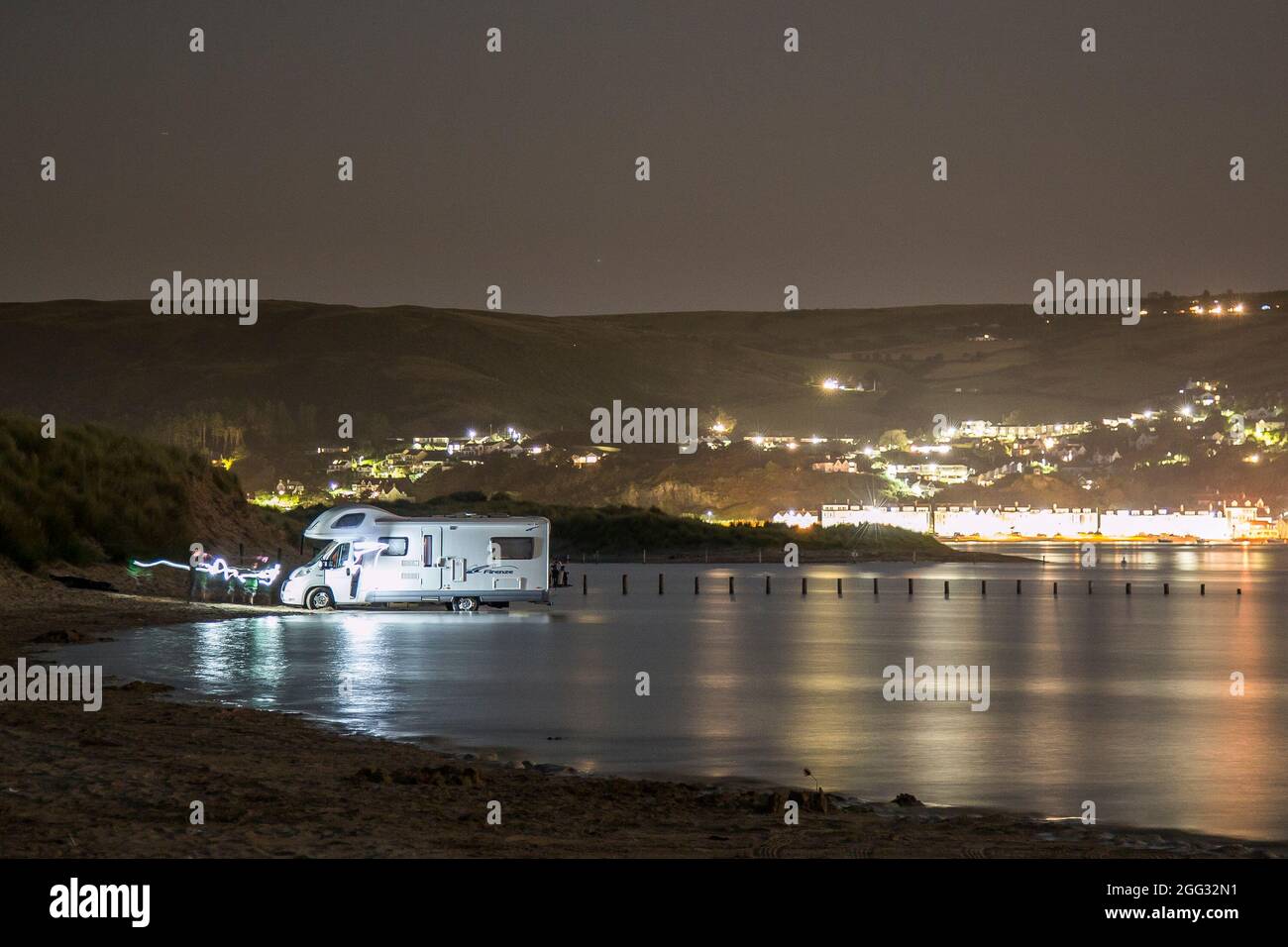 Borth, Regno Unito. 25 agosto 2021. Tempo del Regno Unito: Dopo un'altra giornata calda sulla costa del Galles, i visitatori della spiaggia tornano al loro camper parcheggiato in tarda serata per ritrovarsi bloccati nell'acqua di mare, poiché l'alta marea sembra strisciare più in alto del normale sulle sabbie dell'estuario a Ynyslas, Borth. Cambiamento climatico o solo sfortuna? Le luci di Aberdovey bruciano brillantemente dietro il motorhome attraverso l'estuario, mentre questi villeggianti guardano l'acqua marea in aumento in completo orrore. Credit: Lee Hudson/Alamy Live News Foto Stock