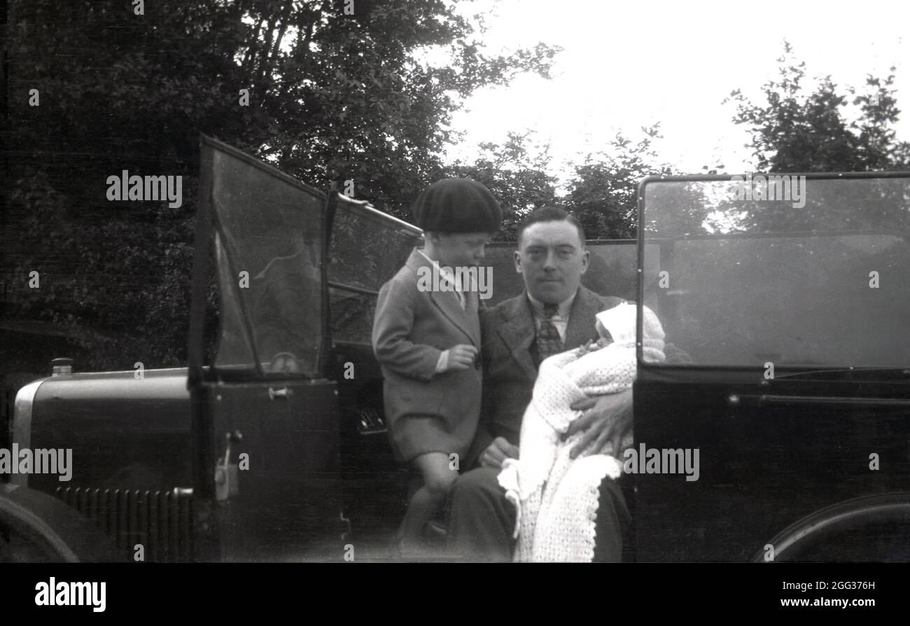 Circa 1930, storico, un giovane ragazzo con suo padre in un'auto a cielo aperto dell'epoca, seduto nel sedile passeggero che tiene un bambino avvolto in una scialle, Inghilterra, Regno Unito. Il ragazzino indossa un cappello, una bacca. Foto Stock