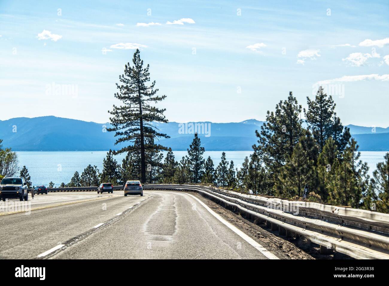 Veicoli che circondano una curva con una barriera di guardia martoriata vicino al lago Tahloe CA Stati Uniti d'America con lago e montagne in lontananza. Foto Stock