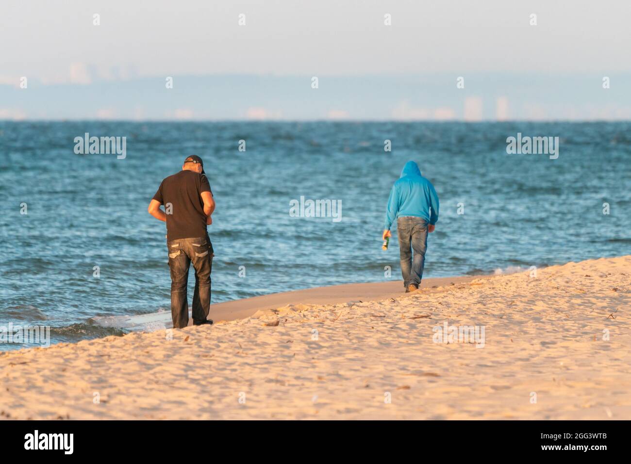 Due uomini che camminano su una spiaggia dal Mar Baltico lontano dallo spettatore all'inizio della mattina d'estate, con la riva nella foschia all'orizzonte lontano. Dopo la festa sensazione. Foto Stock