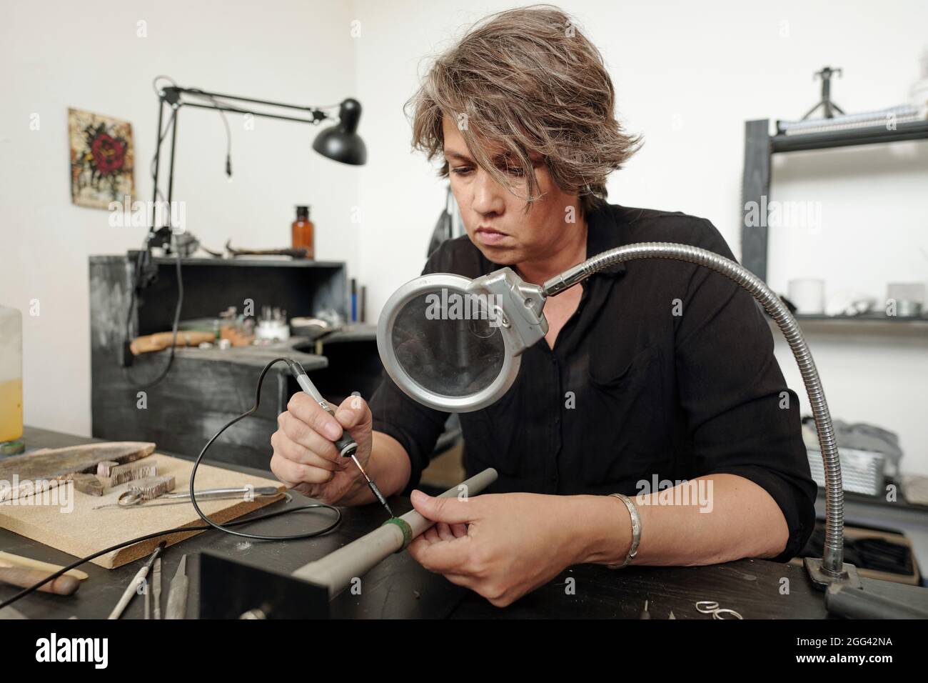 Gioielleria femminile concentrata seduta in officina con mobili neri e pezzi di saldatura insieme sotto lente d'ingrandimento Foto Stock