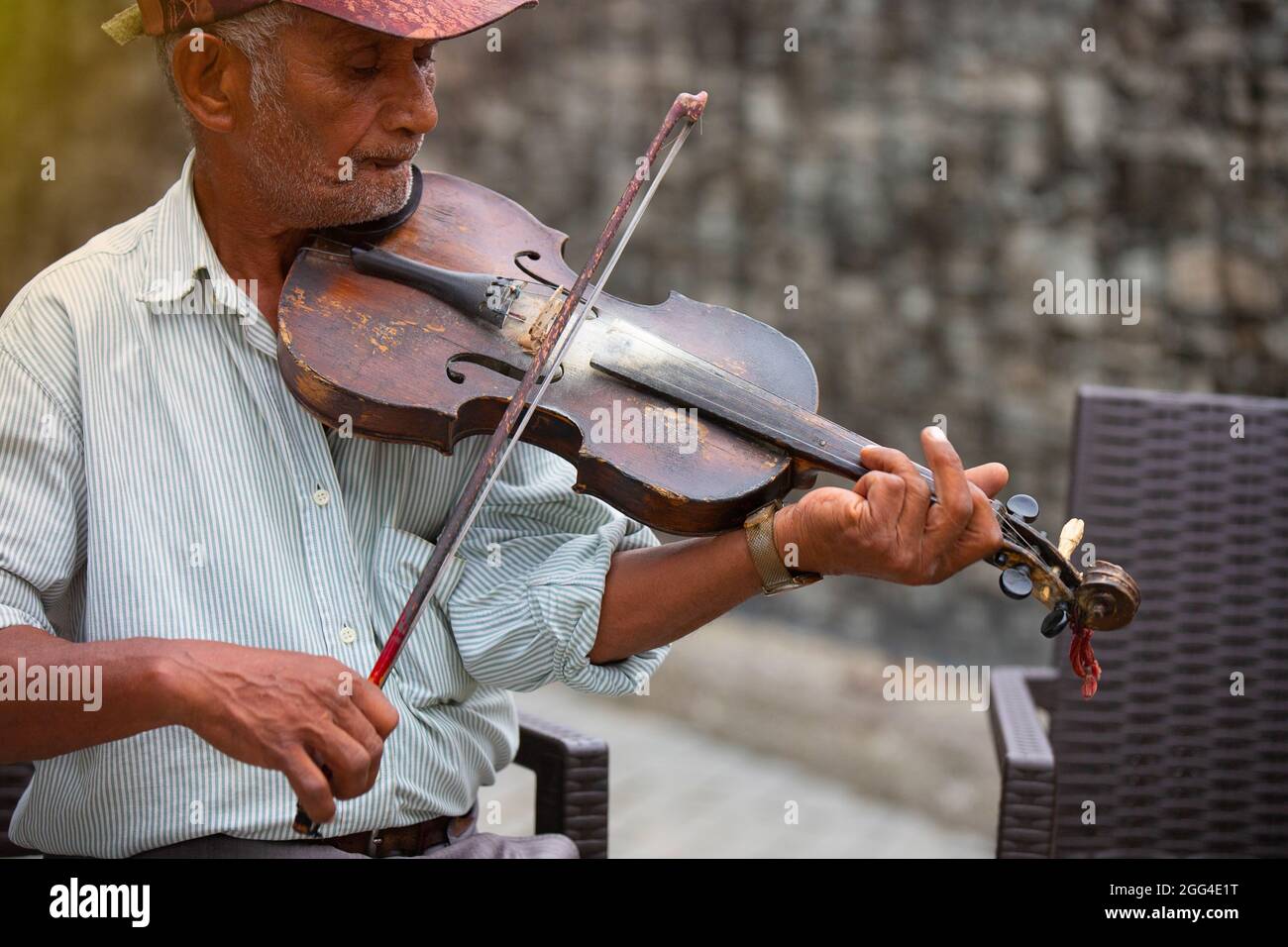 Vecchio musicista di strada seduto su una sedia con un cappuccio sulla testa indossando una t-shirt a righe, suonando violino per tutte le persone che passano accanto a lui e ascoltano la sua Foto Stock