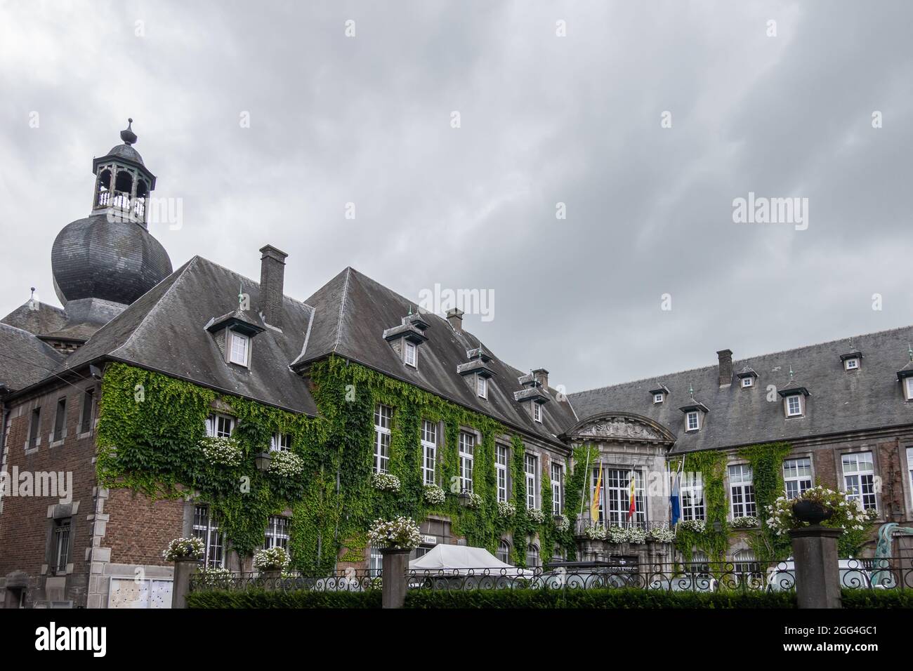 Dinant, Vallonia, Belgio - 8 agosto 2021: Tetto in tegole nere con torre del municipio storico sotto il cielo piovoso. Verde fogliame sale wal Foto Stock